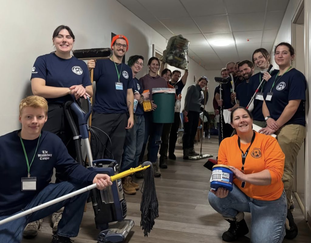 A group of 15 people posing for a photo while holding mops, a vacuum, brooms, cleaning supplies, a trash can, a garbage bag, a roll of paper towels and a container of coffee grounds.