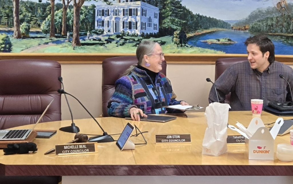 A woman and man are seated at a table, engaged in conversation. City Councilor nametags, Dunkin’ Donuts foodstuffs, laptops, and notepads are spread out around the table.