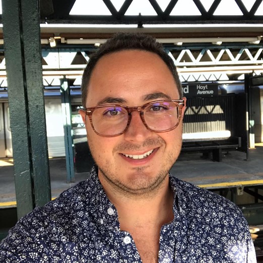 Headshot of James. Wears glasses with a blue print shirt. Smiling.