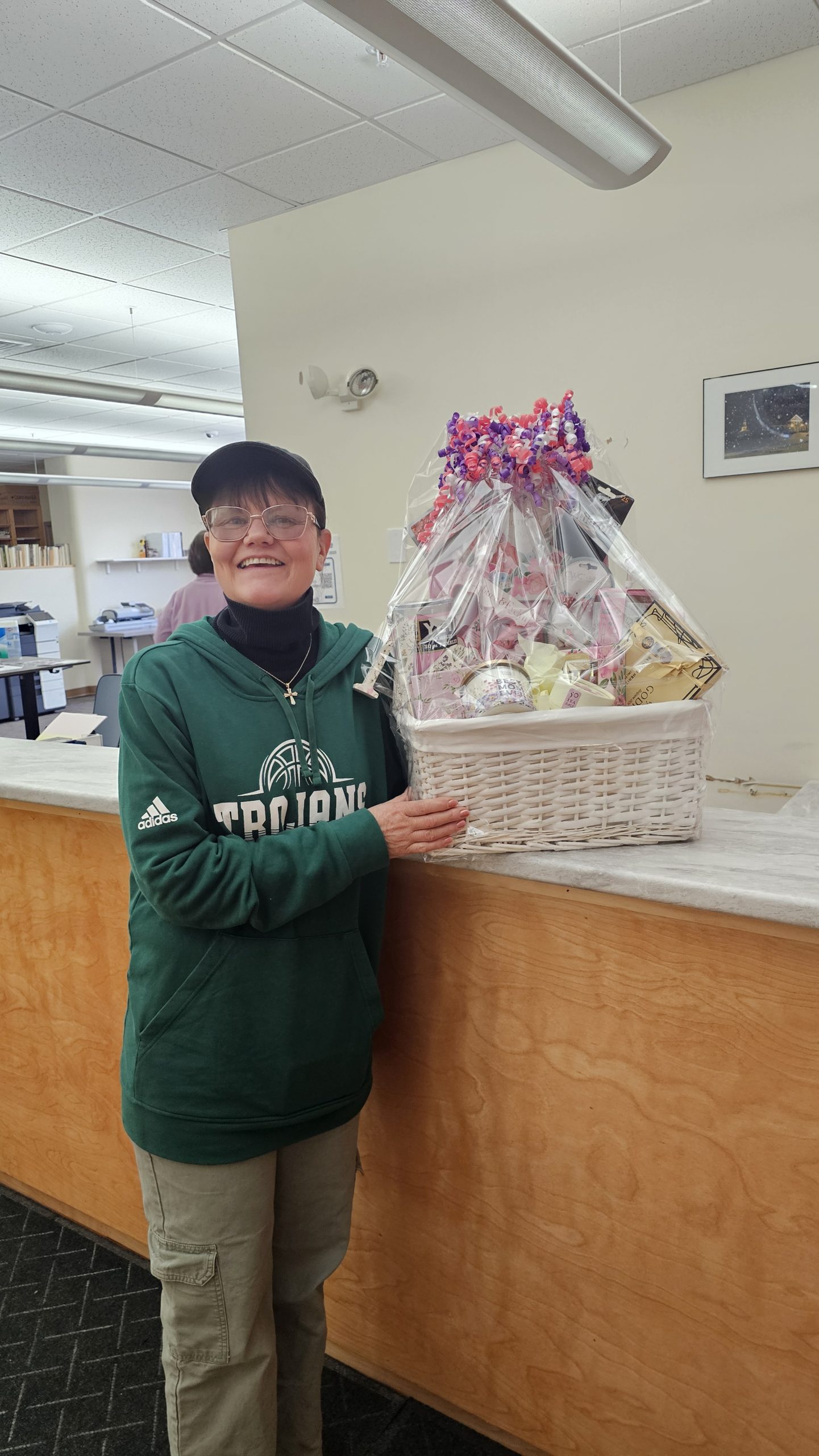 a woman stands next to a gift basket with mother's day themed items. she is smiling and in an office.